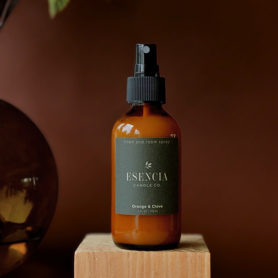 An amber glass room spray bottle with a black spray top and a dark green label reading 'Esencia Candle Co. Orange & Clove', displayed on a wooden block against a warm brown background.