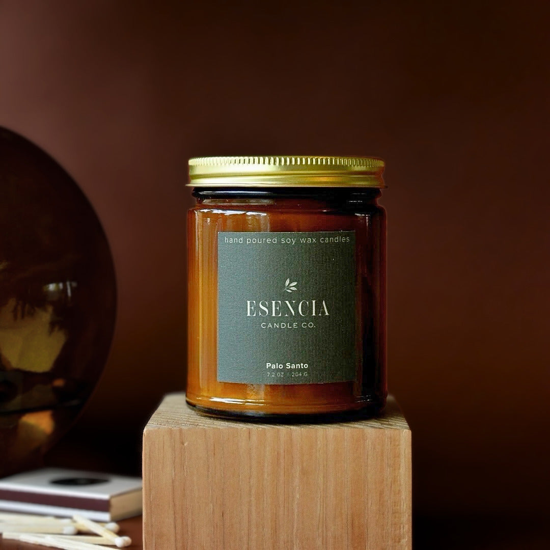 A jar candle from Esencia Candle Co. labeled 'Palo Santo' sits on a wooden block against a dark brown background.
