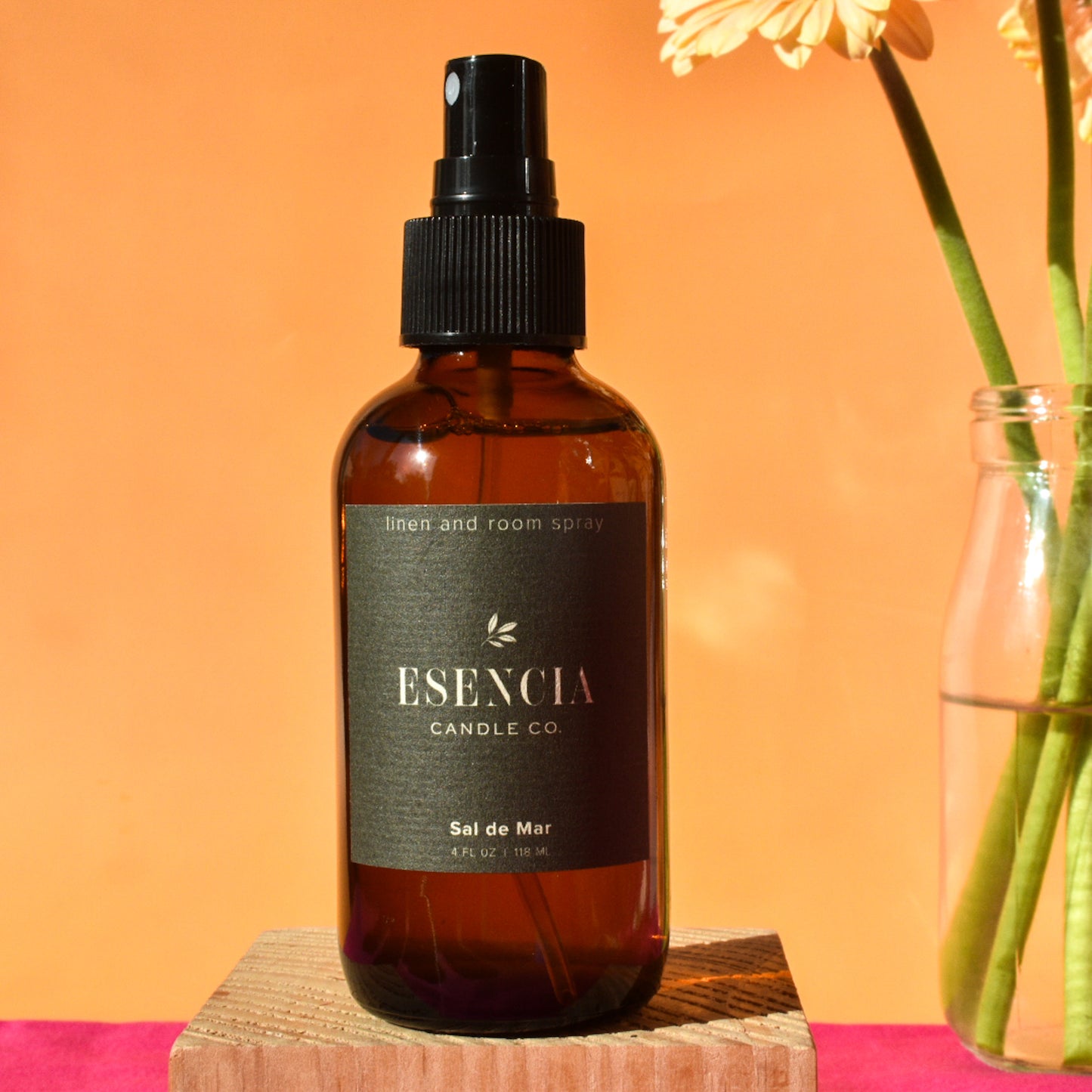 Brown bottle of Esecncia Candle Co. linen and room spray on a wooden block with an orange background.