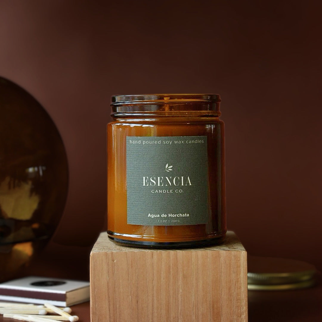 An amber glass jar candle with a dark green label reading 'ESNCIA Candle Co.' sits on a wooden block against a warm brown background.
