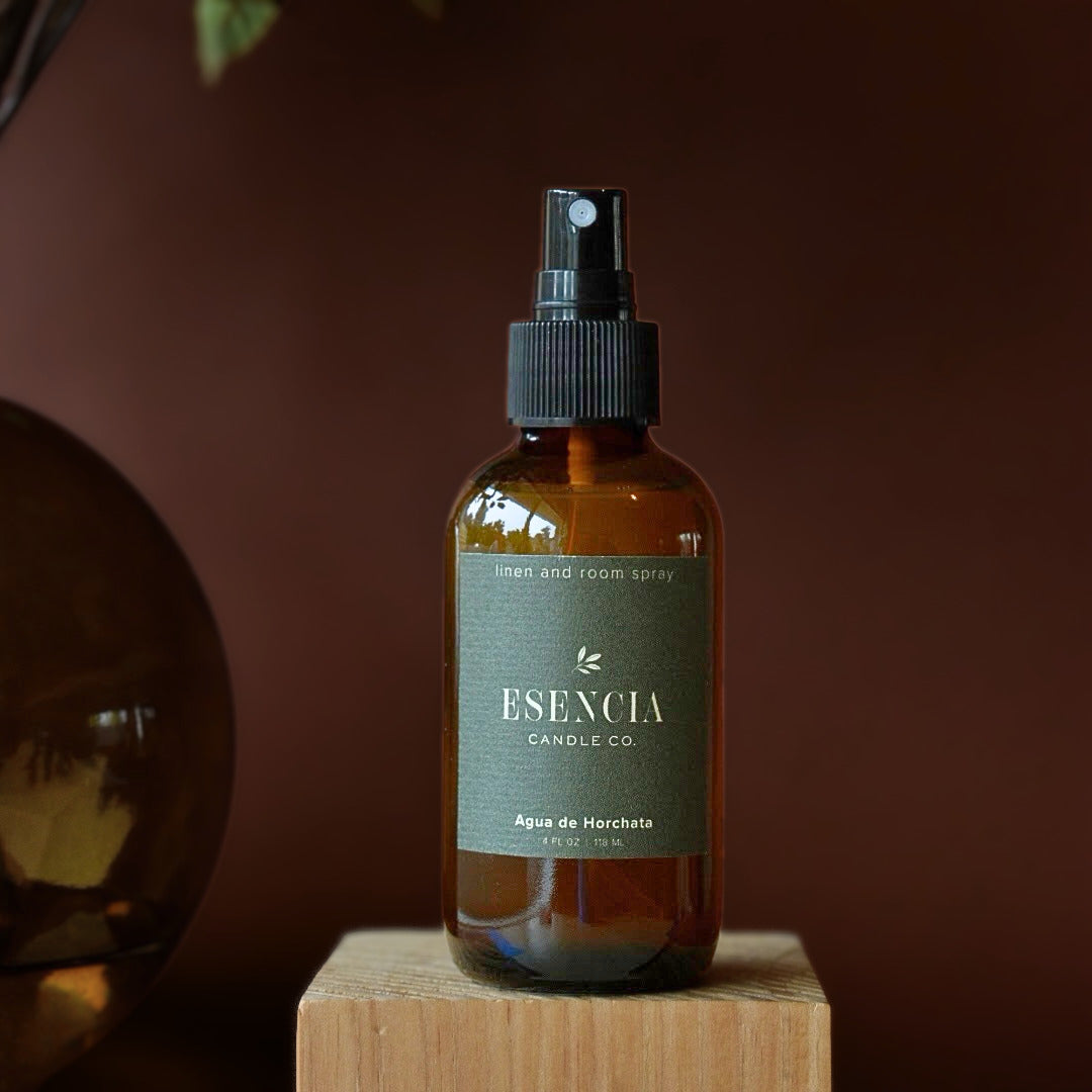 A bottle of ESENCIA Candle Co. linen and room spray in 'Agua de Horchata' scent, featuring an amber glass bottle with a black spray nozzle and a muted green label, displayed on a wooden block against a dark brown background.