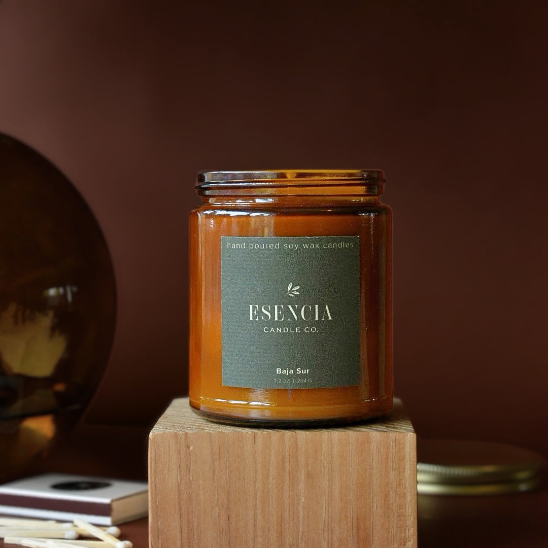 An amber glass jar candle with a dark green label reading 'ESENCIA CANDLE CO. Bajio Sur' sits on a wooden block against a warm brown background.