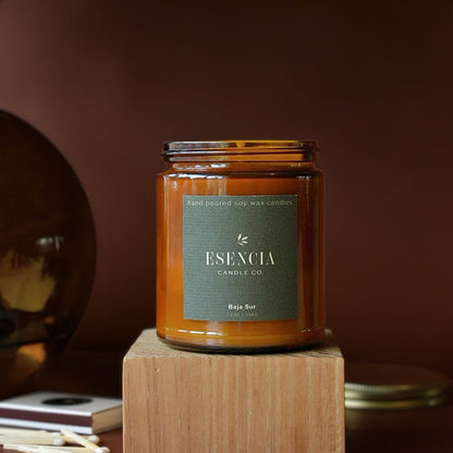 An amber glass jar candle with a dark green label reading 'ESENCIA CANDLE CO. Bajio Sur' sits on a wooden block against a warm brown background.