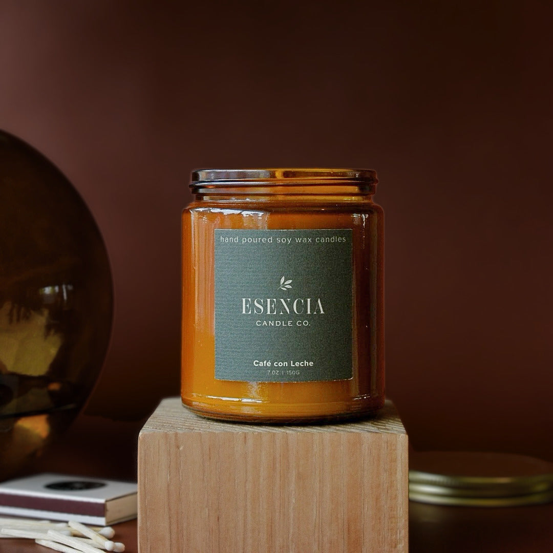 An amber glass jar candle with a dark green label reading 'ESENCIA CANDLE CO.' and 'Café con Leche' sits on a wooden block against a warm brown background.