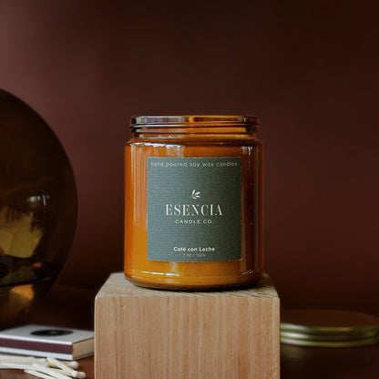 An amber glass jar candle with a dark green label reading 'ESENCIA CANDLE CO.' and 'Café con Leche' sits on a wooden block against a warm brown background.