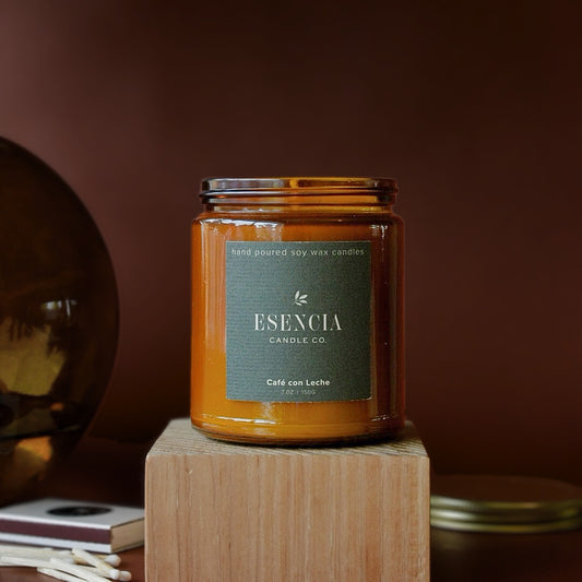 An amber glass jar candle with a dark green label reading 'ESENCIA CANDLE CO.' and 'Café con Leche' sits on a wooden block against a warm brown background.
