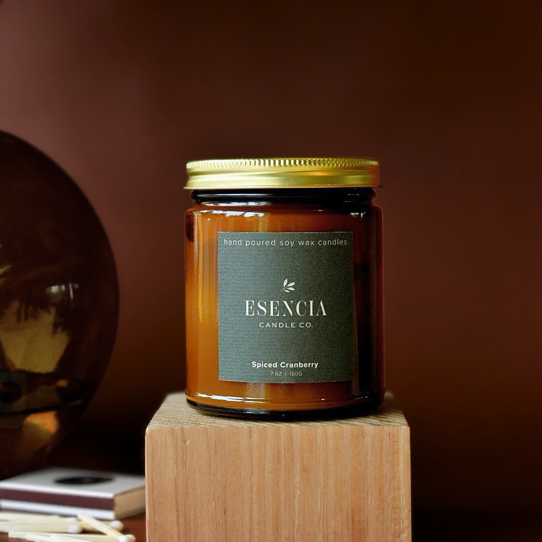 An amber glass jar candle with a gold metal lid and a dark green label reading 'Esencia Candle Co. Spiced Cranberry', displayed on a wooden block against a dark brown background.