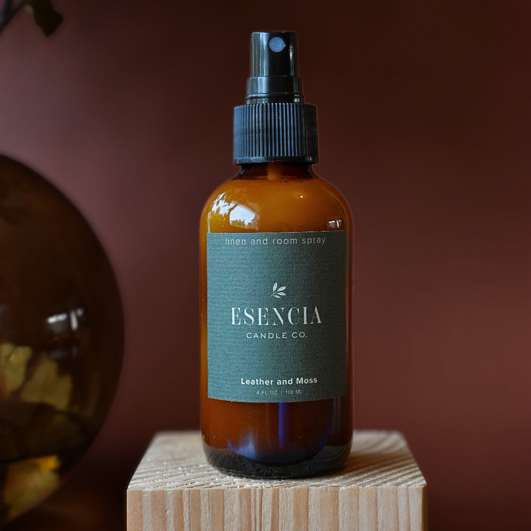 An amber glass room spray bottle with a black spray top and a dark green label reading 'ESENCIA CANDLE CO. Leather and Moss', displayed on a wooden block against a dark brown background.