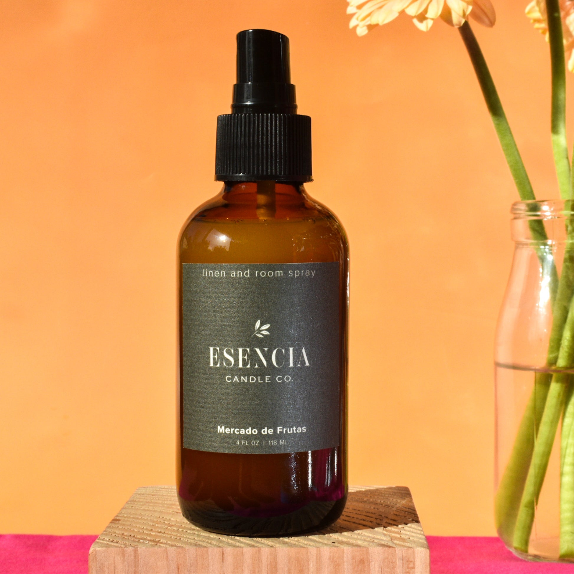 Brown spray bottle with black cap labeled 'Esencia Candle Co' on a pink surface with an orange background.