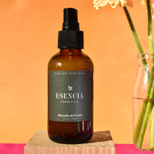 Brown spray bottle with black cap labeled 'Esencia Candle Co' on a pink surface with an orange background.