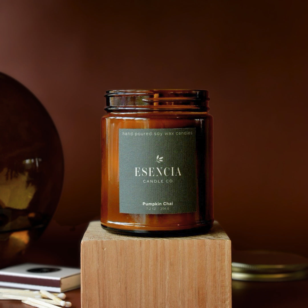 An amber glass jar candle from ESENCIA Candle Co. labeled 'Pumpkin Chai' sits on a wooden block against a warm brown background.