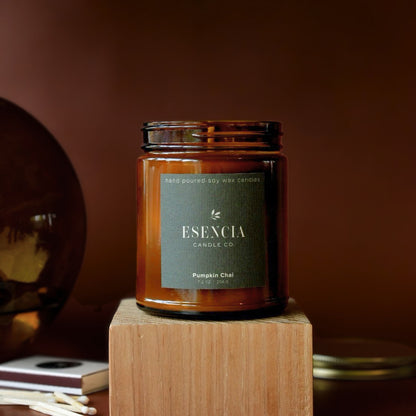 An amber glass jar candle from ESENCIA Candle Co. labeled 'Pumpkin Chai' sits on a wooden block against a warm brown background.