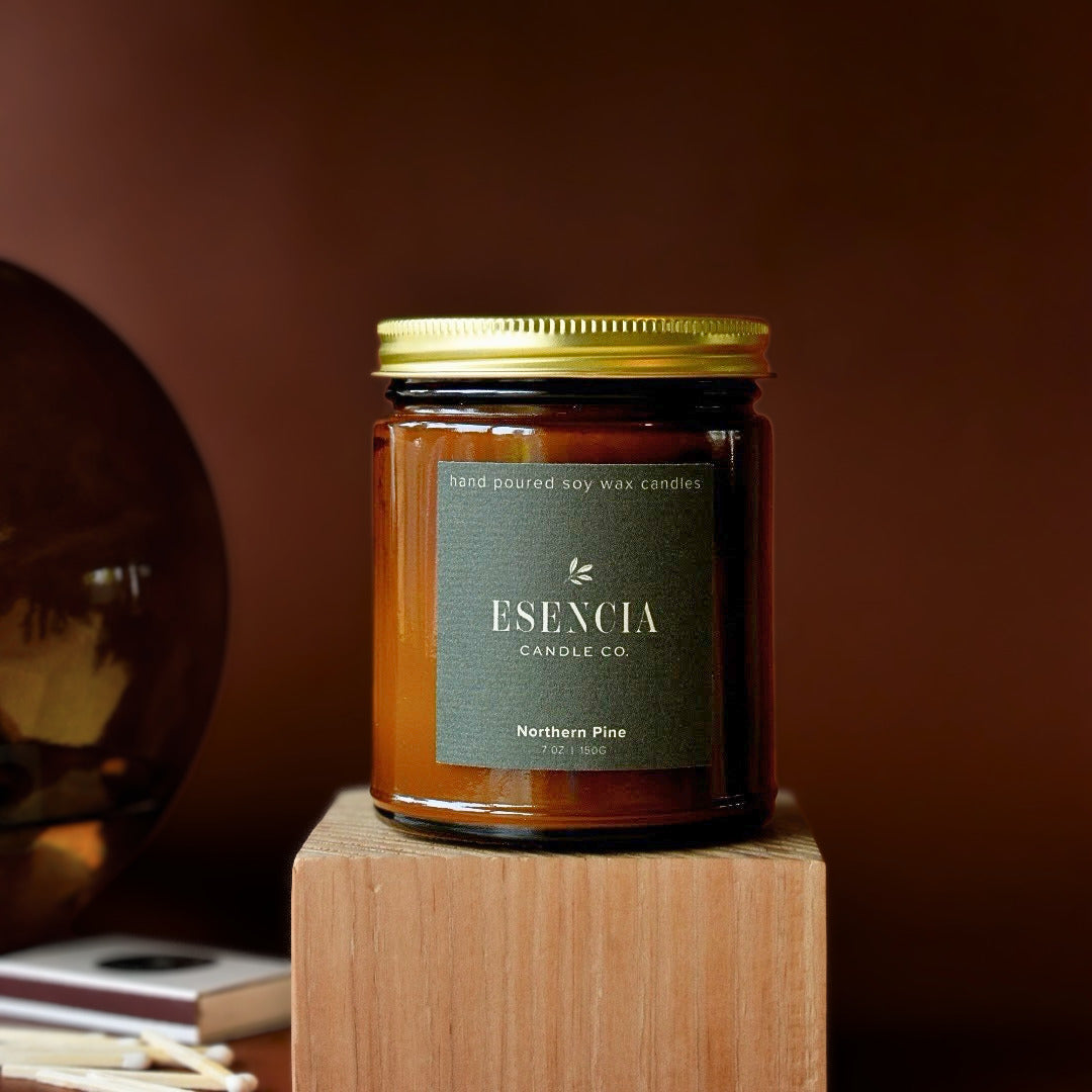 An amber glass jar candle with a gold metal lid and a dark green label reading 'Esencia Candle Co. Northern Pine', displayed on a wooden block against a warm brown background.