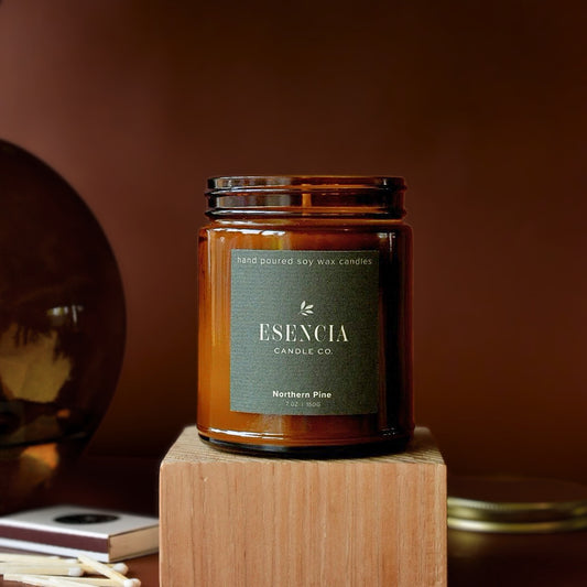 An amber glass jar candle from ESENCIA Candle Co. sits on a wooden block against a warm brown background.