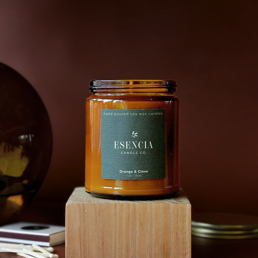An amber glass jar candle from ESENCIA Candle Co. on a wooden block, set against a dark brown background with a decorative mirror and books blurred in the foreground.