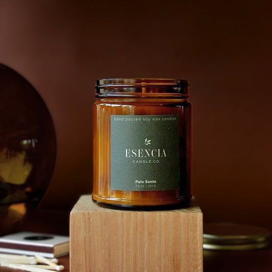 An amber glass jar candle with a dark green label reading 'ESENCIA CANDLE CO.' and 'Palo Santo,' displayed on a wooden block against a warm brown background.