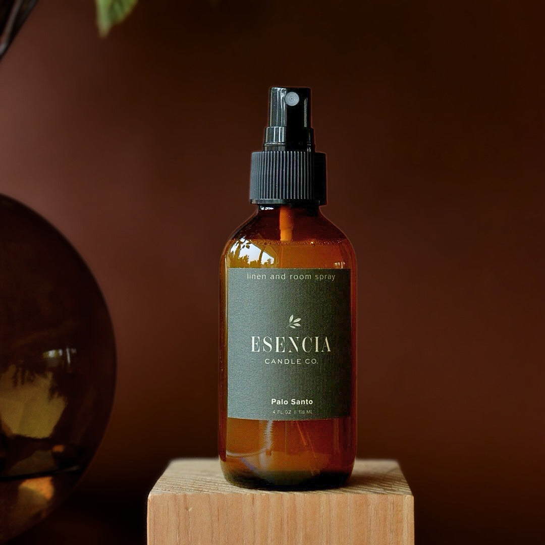 A bottle of ESENCIA Candle Co. linen and room spray in the 'Pale Sage' scent, displayed on a wooden block against a warm brown background.
