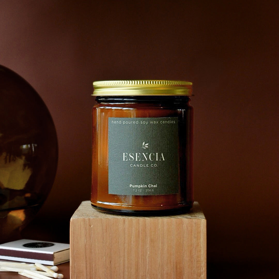 A glass jar candle with amber-colored wax and a gold metal lid, standing on a wooden block against a dark brown background. The front label reads 'ESENCIA CANDLE CO.