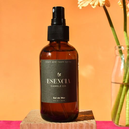 Brown bottle of Esecncia Candle Co. linen and room spray on a wooden block with an orange background.