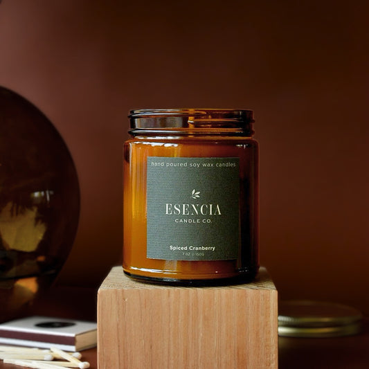 An amber glass jar candle labeled 'ESENCIA Candle Co. Spiced Cranberry' sitting on a wooden block against a warm brown background.